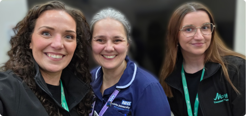 Three smiling women taking a selfie together; the woman in the center is wearing a blue NHS uniform.
