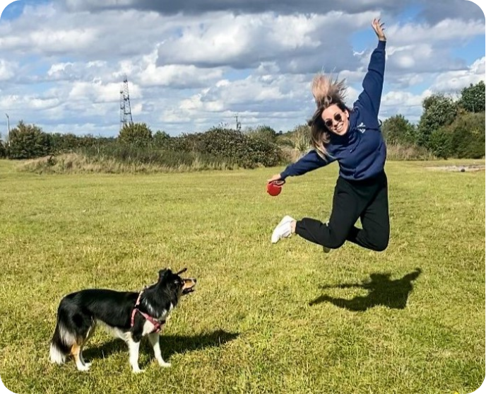 A woman jumps joyfully in an open field while her dog looks on.
