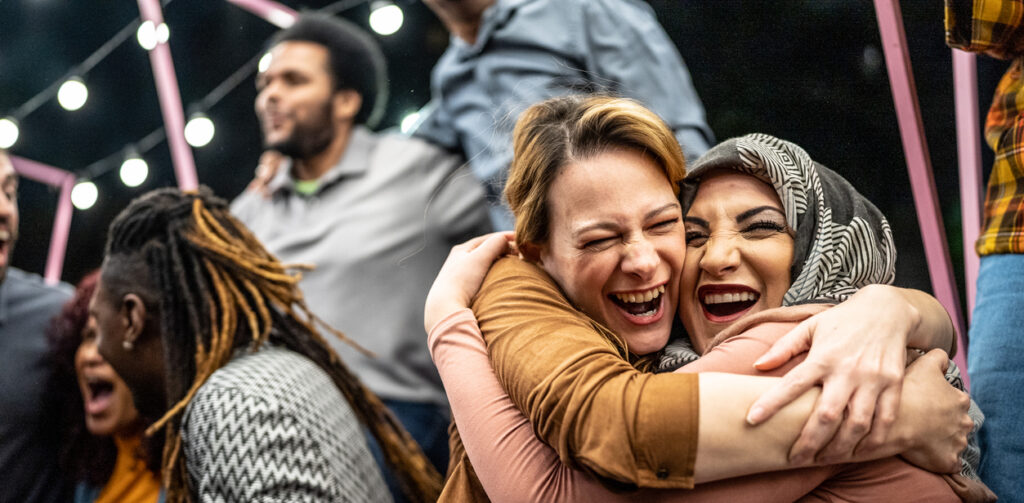 Two women laugh joyfully while sharing a close hug at an outdoor evening gathering.