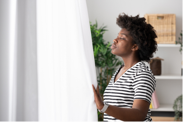 A Black woman standing by a bright window with sheer white curtains