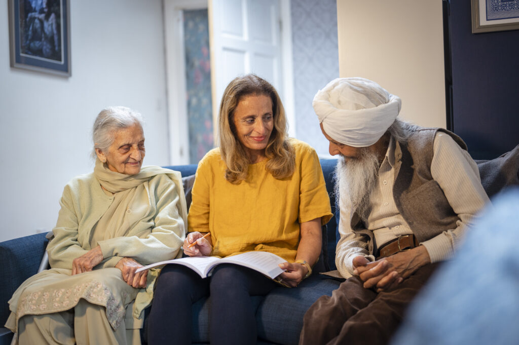 A smiling woman sits on a sofa between two elderly adults, including a man wearing a turban, showing them an open book.
