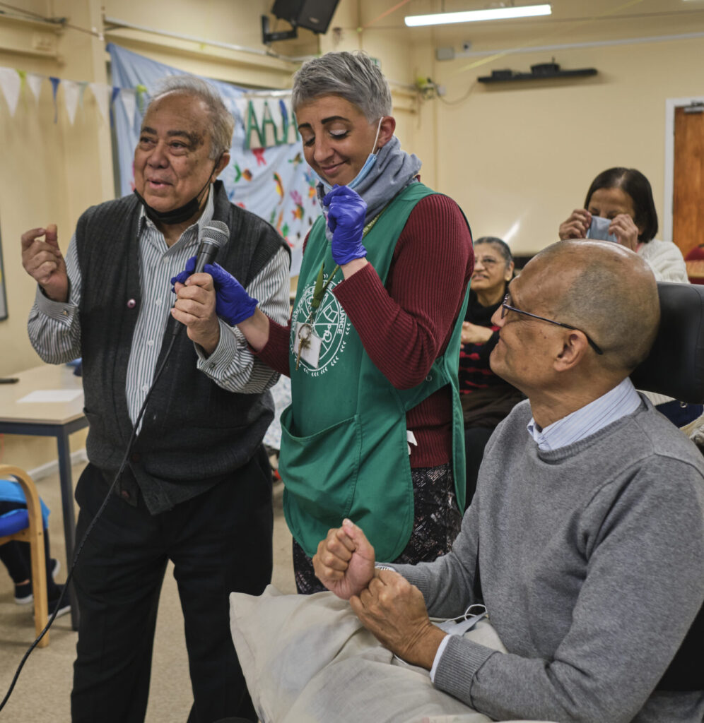 An older man holding a microphone and a woman in a green apron interact with a smiling man seated in a wheelchair.