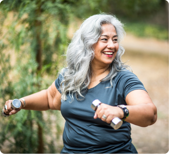 A smiling older woman exercising outdoors with small dumbbells.
