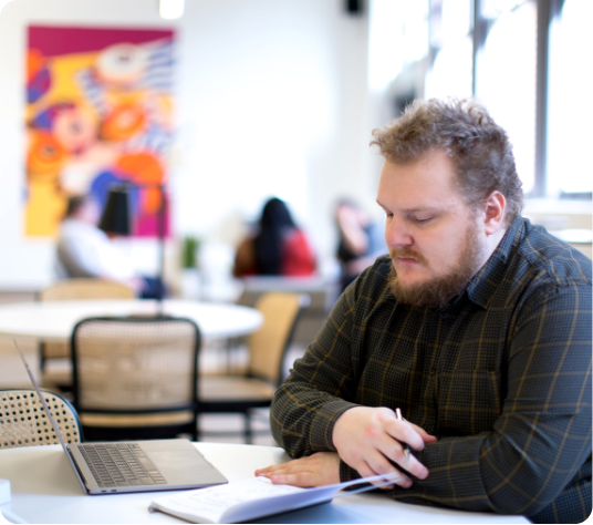 A bearded man focused on writing in a notebook next to his laptop.