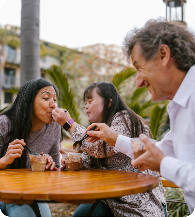 A woman, a girl with Down syndrome, and a man are sitting at a table outdoors, eating from small containers with spoons.