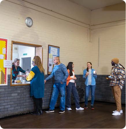 A diverse group of adults queuing and socializing at a beverage counter in a community hall.