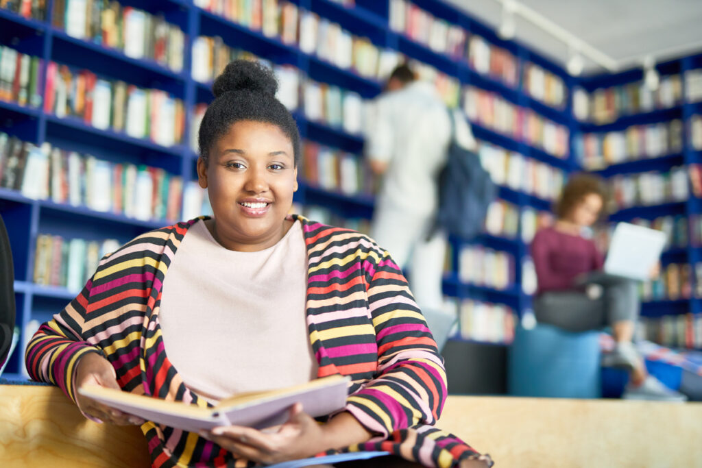 Smiling Black teenage girl holding open book in library with tall blue bookshelves.