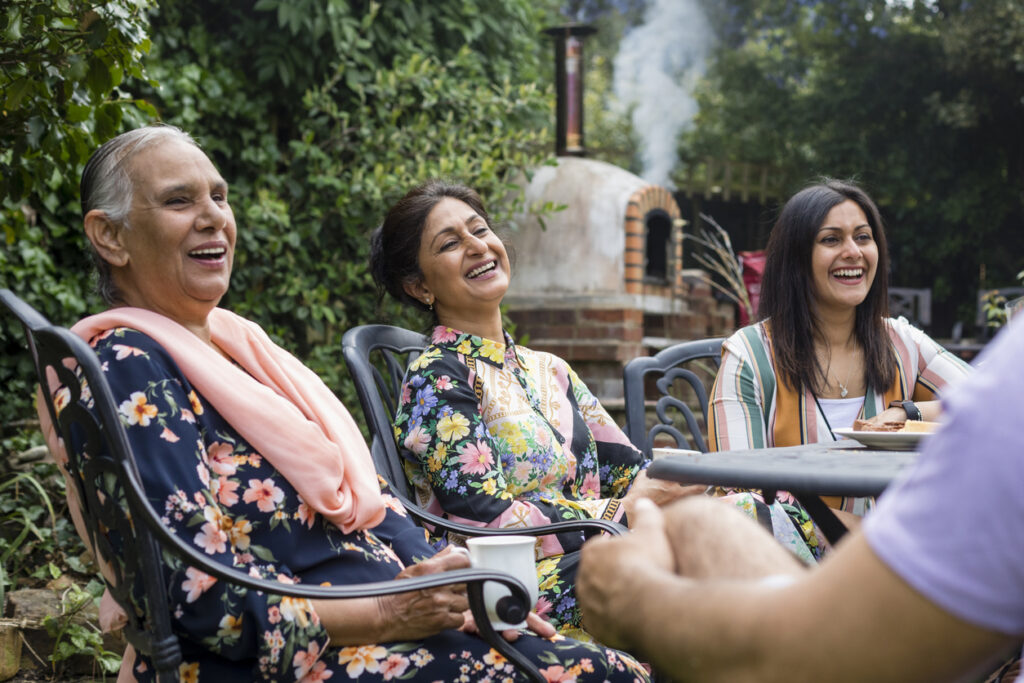 Three women laughing together outdoors in a garden during a social gathering with friends nearby.